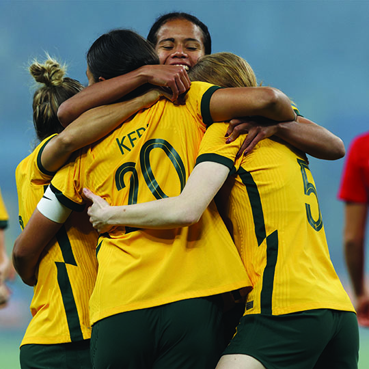 A group of four Australian football athletes gather in a group hug, wearing their green and gold uniforms